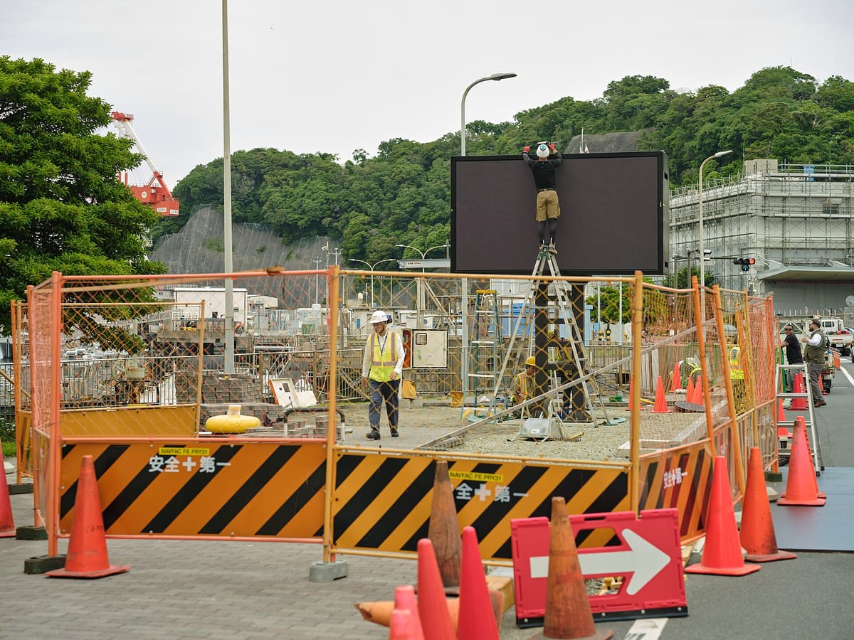 Technician on a ladder mounting an outdoor LED sign inside a fenced installation site